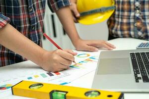 Asian engineer and partner team learn and talk with house plan project blueprint, graph and yellow helmet and engineering construction tools on table work. photo