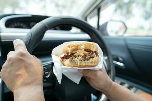 asiático dama participación hamburguesa a comer en auto, peligroso y riesgo un accidente. foto