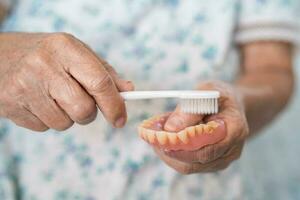 asiático mayor mujer paciente utilizar cepillo de dientes a limpiar parcial dentadura de reemplazo dientes. foto