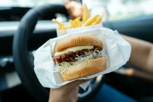 asiático dama participación hamburguesa y francés papas fritas a comer en auto, peligroso y riesgo un accidente. foto