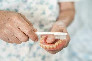 asiático mayor mujer paciente utilizar cepillo de dientes a limpiar parcial dentadura de reemplazo dientes. foto