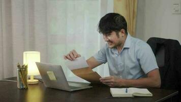 asiático hombre recibe contento Noticias en el correo. en el hogar, él se sienta a un mesa con un computadora portátil, sostiene un sobre en su manos, lee, sonrisas video