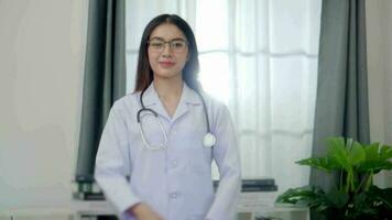 ásia mulher médico em pé com braços cruzado é vestindo uniforme branco roupão e estetoscópio dentro exame quarto às a clínica. video