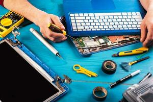 el asistente repara la computadora portátil con herramientas y manos en la mesa de madera azul. vista superior foto