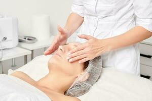 Cosmetologist rubbing moisturizing cream mask into woman face skin for rejuvenation in beauty salon photo