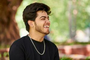 Smiling young indian man portrait in black t shirt and silver neck chain outdoor green public park photo