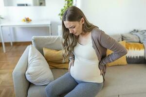 Young pregnant woman suffering from backpain or migraine, feeling sick, sitting on sofa at home, free space. Sad expecting lady touching her big tummy, home alone photo