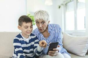 Happy grandmother and grandson using phone together, sitting on cozy sofa at home, browsing mobile device apps, grandma with grandchild playing game, looking at screen, having fun photo