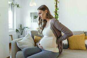 Pregnant woman suffering during her pregnancy, with back pain and headaches. Pregnant woman sitting on the sofa holding her belly with worried face expression. photo
