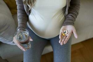 Healthcare concept. Cropped view of pregnant woman take vitamin pill, holding cup with water in hands, sitting alone behind wooden table photo