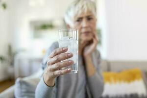 Senior woman with sensitive teeth and hand holding glass of cold water with ice. Healthcare concept. Mature woman drinking cold drink, glass full of ice cubes and feels toothache, pain photo