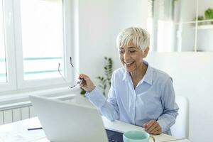 Focused old woman with white hair at home using laptop. Senior stylish entrepreneur working on computer at home. Woman analyzing and managing domestic bills and home finance photo