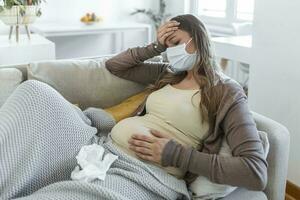 pregnant young adult woman resting on sofa at home, feeling unwell. Young pregnant woman has suffered from covid-19 sitting on the sofa with face mask. Pregnancy symptoms photo