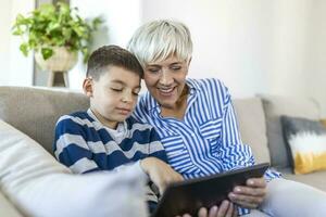 Happy grandmother and grandson using tablet together, sitting on cozy sofa at home, browsing tablet device apps, grandma with grandchild playing game, looking at screen, having fun photo