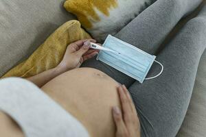 Young pregnant woman lying in bed, measuring body temperature, holding thermometer and face mask photo