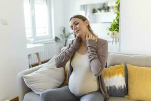 pregnant woman suffer neck and shoulder pain. pregnant woman holding her injured neck while touching her belly and sitting on couch photo