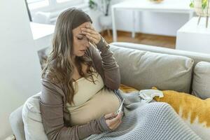 Young pregnant woman lying in bed, measuring body temperature, holding thermometer feeling sick photo