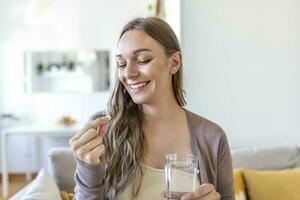 Head shot portrait happy woman holds pill glass of water, takes daily medicine vitamin D, omega 3 supplements, skin hair nail strengthen and beauty, medication for health care concept photo