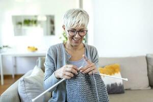 Elderly woman in glasses sit on couch at home smile holding knitting needles and yarn knits clothes for loved ones, favorite activity and pastime, retired tranquil carefree life concept photo