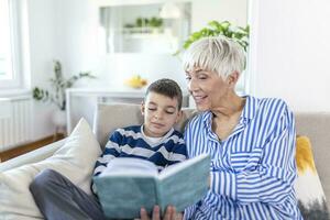 amoroso abuela enseñando nieto participación libro sentado en sofá, abuela bebé modelo abrazando niño chico leyendo cuento de hadas a linda niño, niñera abuelita narración historia a preescolar nieto foto