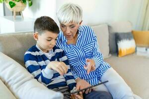 Happy grandmother and grandson using tablet together, sitting on cozy sofa at home, browsing tablet device apps, grandma with grandchild playing game, looking at screen, having fun photo