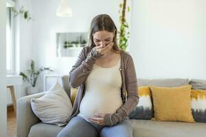 young pregnant woman suffering from toxicosis at home. maternity lady having morning sickness covering mouth with hands while sitting on couch in living room. mom expect unborn baby photo