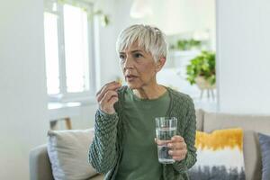Senior woman takes pill with glass of water in hand. Stressed female drinking sedated antidepressant meds. Woman feels depressed, taking drugs. Medicines at work photo