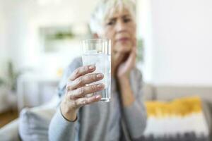 Senior woman with sensitive teeth and hand holding glass of cold water with ice. Healthcare concept. Mature woman drinking cold drink, glass full of ice cubes and feels toothache, pain photo