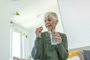 Senior woman takes pill with glass of water in hand. Stressed female drinking sedated antidepressant meds. Woman feels depressed, taking drugs. Medicines at work photo