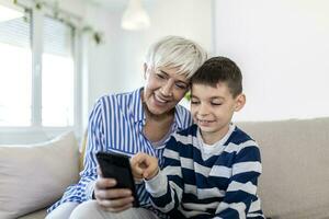 Happy grandmother and grandson using tablet together, sitting on cozy sofa at home, browsing tablet device apps, grandma with grandchild playing game, looking at screen, having fun photo