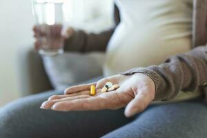 Healthcare concept. Cropped view of pregnant woman take vitamin pill, holding cup with water in hands, sitting alone behind wooden table photo