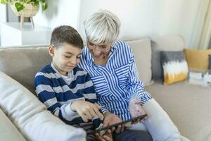 Happy grandmother and grandson using tablet together, sitting on cozy sofa at home, browsing tablet device apps, grandma with grandchild playing game, looking at screen, having fun photo