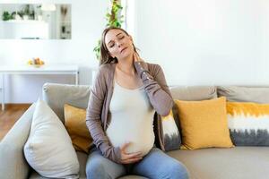 pregnant woman suffer neck and shoulder pain. pregnant woman holding her injured neck while touching her belly and sitting on couch photo