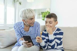 Happy grandmother and grandson using phone together, sitting on cozy sofa at home, browsing mobile device apps, grandma with grandchild playing game, looking at screen, having fun photo