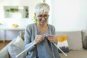 Elderly woman in glasses sit on couch at home smile holding knitting needles and yarn knits clothes for loved ones, favorite activity and pastime, retired tranquil carefree life concept photo