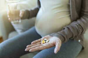 Healthcare concept. Cropped view of pregnant woman take vitamin pill, holding cup with water in hands, sitting alone behind wooden table photo