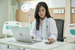 Young beautiful Asian female doctor online working with laptop and smartphone on medical consultation application via internet at working desk in hospital clinic. photo