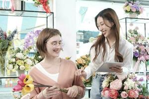 Two young beautiful female florist partners discuss bloom bouquet arrangement design in tablet, online purchase order work in colorful flower shop store with fresh flora, SME business entrepreneur. photo