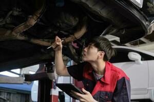 Professional young Asian male motor mechanic inspects undercarriage of electric car lifted by forklift jack for repair at garage, automotive maintenance service works industry occupation business. photo