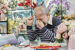 Asian elderly male florist owner is unwell with headaches and health deteriorating, stress from overwork in a flower shop, SME business entrepreneur. photo