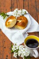 Baked open rolls and a cup of coffee on a dark, worn rustic wooden table. The composition is decorated with a twig with white flowers. Cherry tree flowers. Selective focus. photo