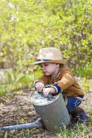 Cute little toddler boy in a hat and rubber boots is watering plants with a watering can in the garden. A charming little kid helping his parents grow vegetables. photo