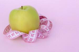 A green apple and a centimeter measuring tape on a pink background. Sports, sports nutrition, healthy eating, diet. Space for the text. photo
