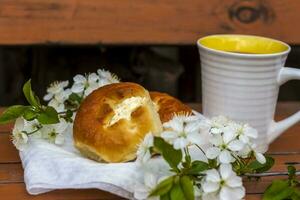 Baked open rolls and a cup of coffee on a dark, worn rustic wooden table. The composition is decorated with a twig with white flowers. Cherry tree flowers. Selective focus. photo