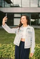 Young woman in white denim jacket taking selfie on street. Office building in the background photo