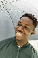 Young African American man under black umbrella in rain, smiling. Fall or spring weather photo