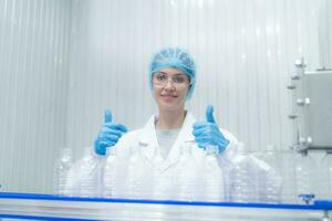 Portrait thumbs up of smiling Caucasian technologist expert standing by an automated machine for PET water production in a bottling factory. photo