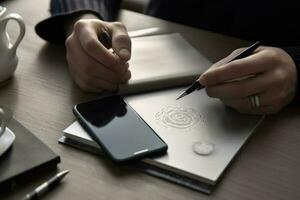 a person, likely a business professional, sitting at a table and writing on a notepad with a pen. Their cell phone is placed nearby on the table. The scene conveys a sense of organization. photo