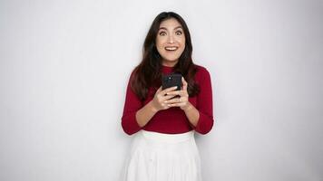 un retrato de un contento asiático mujer vistiendo un rojo camisa mientras participación su teléfono, aislado por blanco antecedentes video