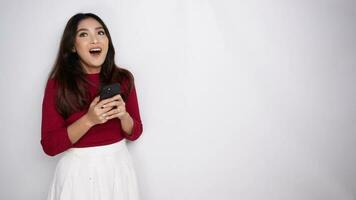 A thoughtful young Asian woman is wearing red t-shirt and holding her phone looks inspired by ideas, isolated by white background video